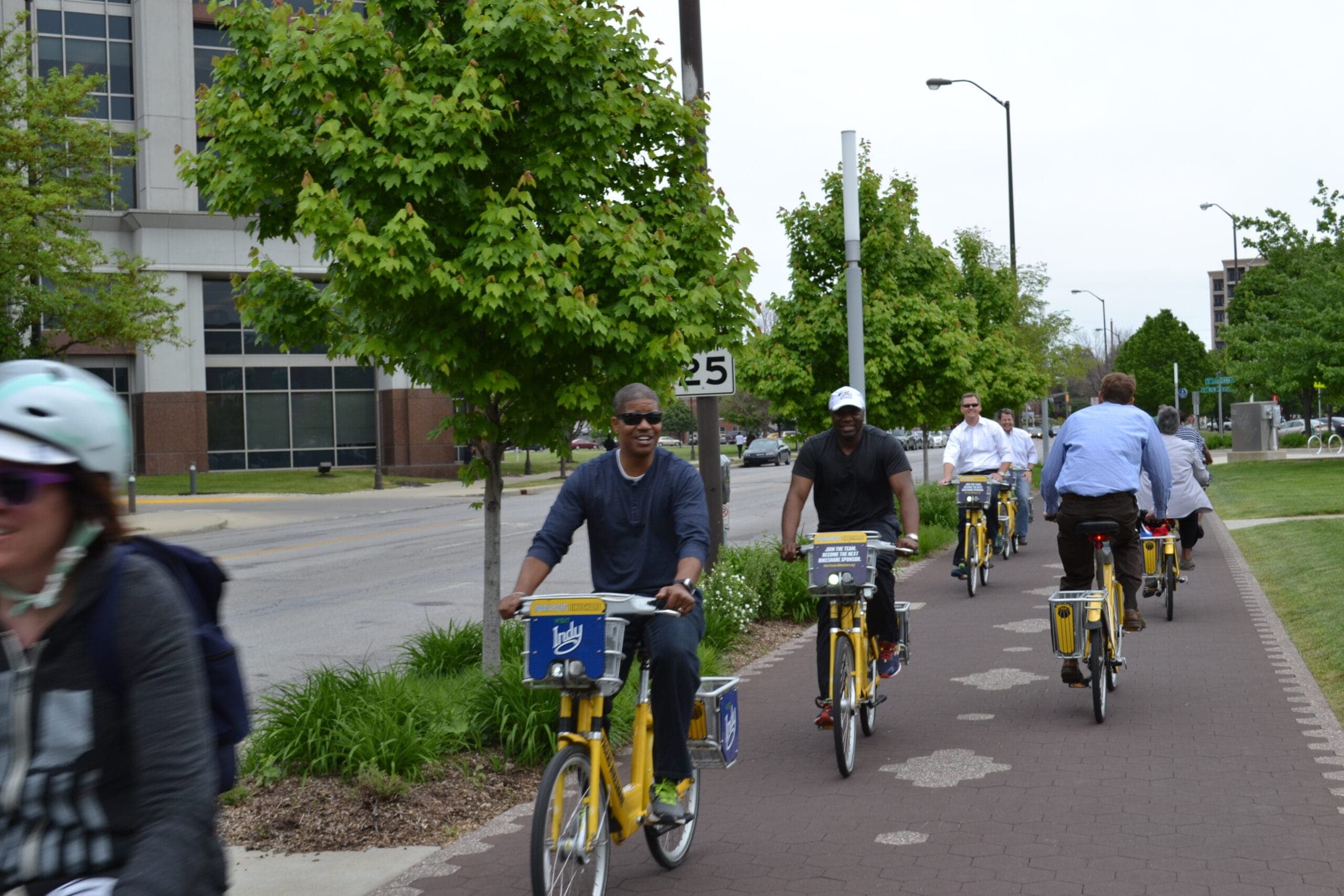 A group of black men ride bikes down a tree-lined path next to a 25 mph street