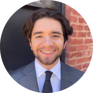 Headshot of Corrigan Salerno, wearing a gray suit jacket and navy tie against a brick and industrial background outside.